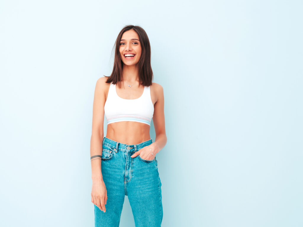Beautiful Smiling Woman Dressed In White Jersey Top Shirt And Jeans. Sexy Carefree Cheerful Model Enjoying Her Morning. Adorable And Positive Female Posing Near Light Blue Wall In Studio