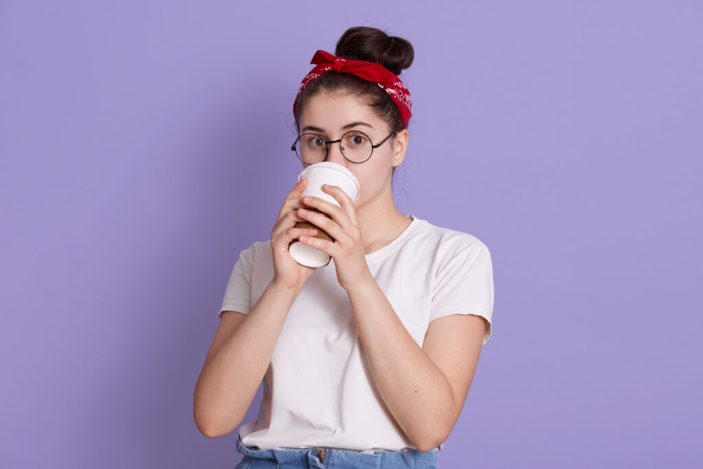 Winsome Young Happy Beautiful Woman In Red Hairband And White Casual T Shirt, Poses Against Lilac Background, Drinking Coffee In Paper Cup, Enjoying Hot Beverage.